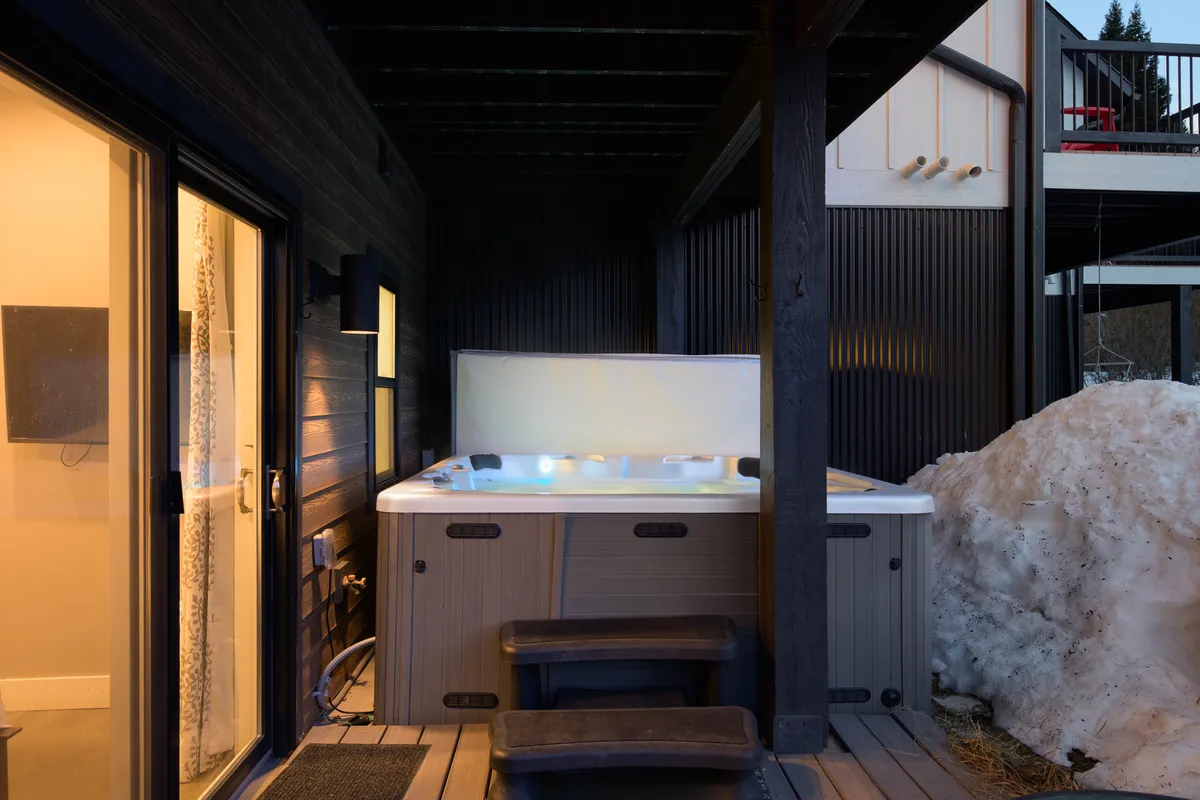 A cozy outdoor hot tub sits steaming beside snow drifts and wooden steps, with people relaxing and enjoying the warm water under a winter night sky, creating an inviting post-adventure retreat.