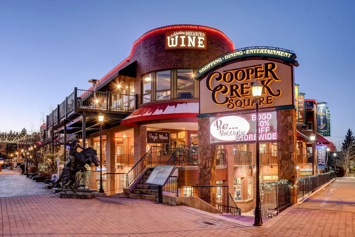 A lively plaza featuring the Cooper Creek Square sign, wine shop signage, a statue of a horse, and inviting outdoor seating areas. The festive setting suggests easy access to pet-friendly patios, local shops, and community events, supporting the narrative of a vibrant, walkable neighborhood.