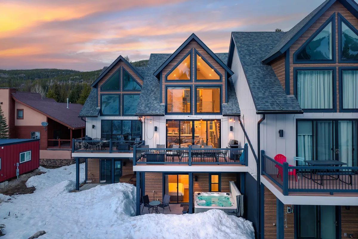 A stunning winter exterior shot of the villa, featuring a large glass façade, an outdoor hot tub, balcony seating, and snow-covered grounds with mountains beyond. The image exudes tranquility and rejuvenation, illustrating the perfect spot for après-adventure relaxation and storytelling.