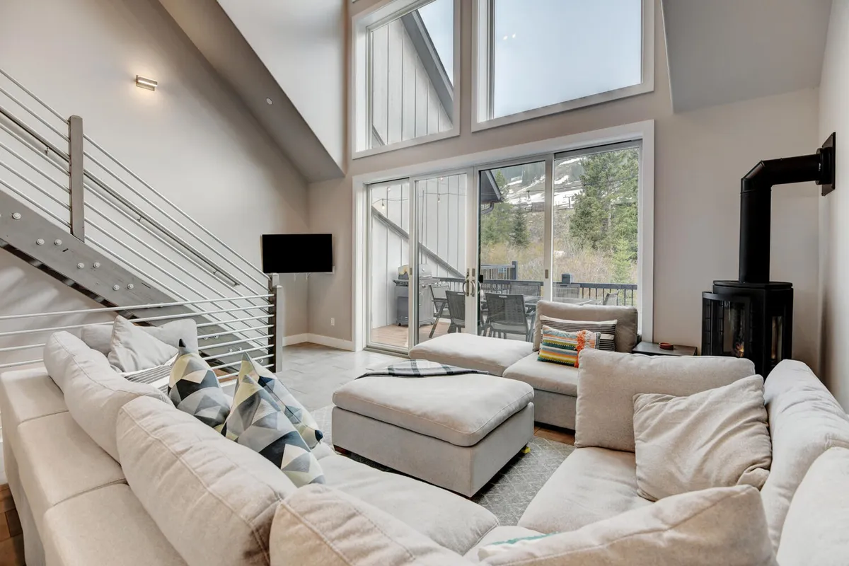 This image captures a spacious great room featuring a large sectional sofa positioned to enjoy both the cozy warmth of a stove and sweeping floor-to-ceiling windows. The windows provide expansive mountain views and fill the space with natural light, while the layout encourages socializing and relaxation.
