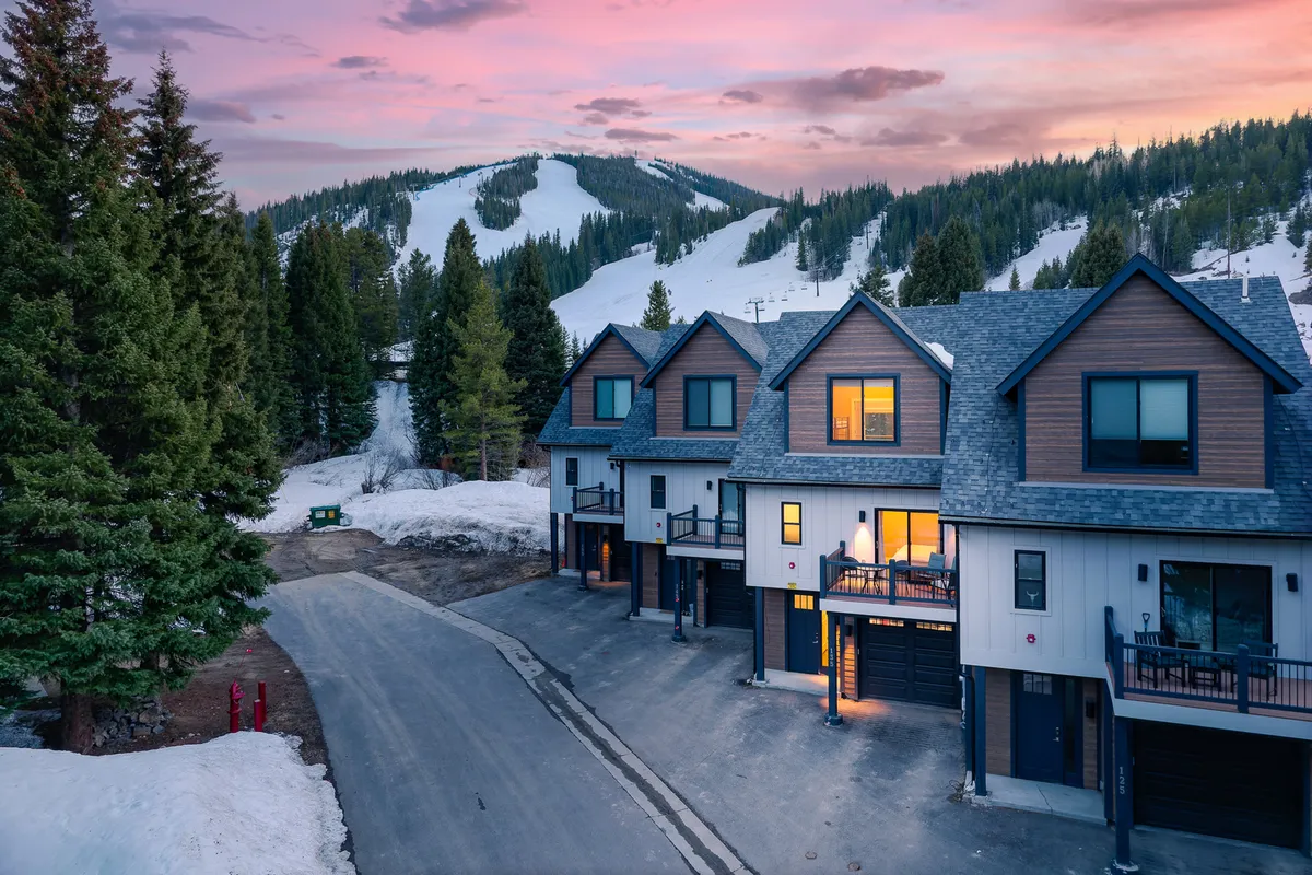 The image features a row of modern townhouses nestled at the base of a mountain slope, with snow-capped peaks in the background and a stunning pink and purple sunset sky overhead. The scene hints at both adventure and relaxation, with inviting porches perfect for unwinding after a day outdoors.