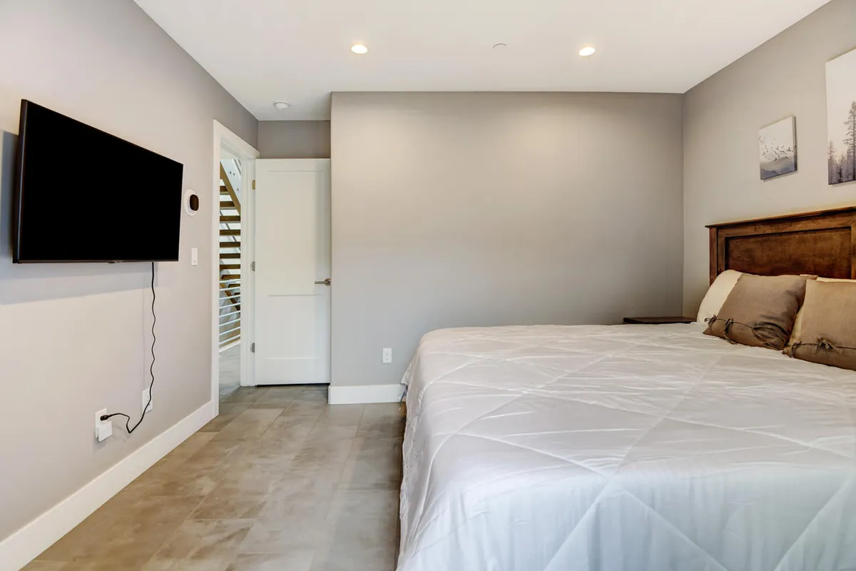 A tranquil bedroom featuring a bed with crisp white bedding, a flat-screen TV mounted on the wall, a wooden headboard, and minimalist decor. Natural light filters in, enhancing the serene ambiance ideal for relaxation or enjoying a quiet movie night.