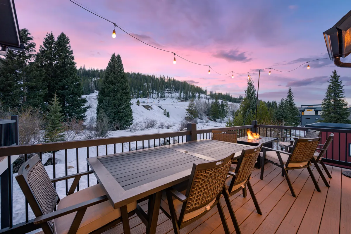 An outdoor seating and dining area set under string lights, with a cozy fire pit and expansive mountain views at sunset. The scene suggests a welcoming space for gathering, relaxing, and enjoying the fresh mountain air after a day of adventure.