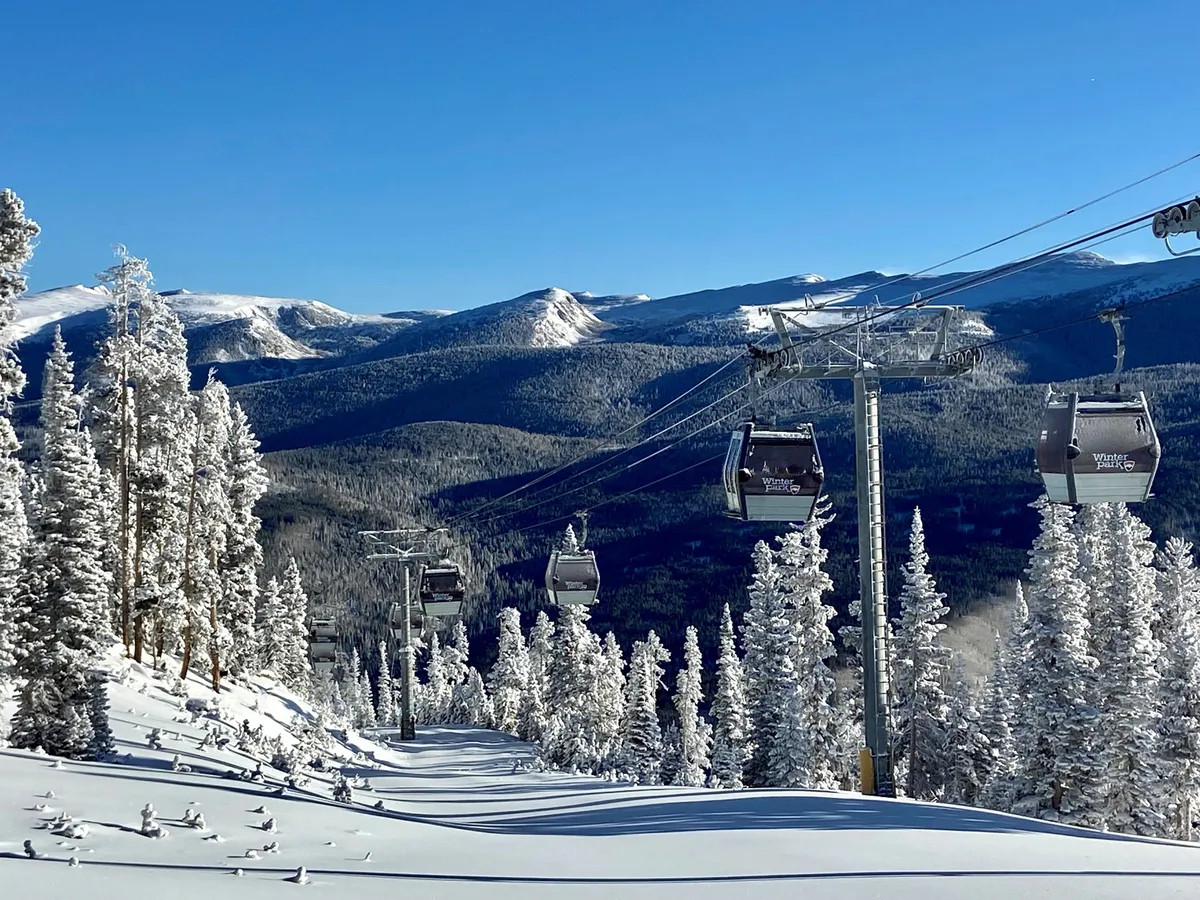 A breathtaking winter scene with ski lift gondolas ascending above snow-covered trees and a magnificent mountain backdrop. The slopes are visibly groomed for skiing and snowboarding, and the entire area is enveloped in fresh snow, conjuring the essence of a ski resort in full swing.