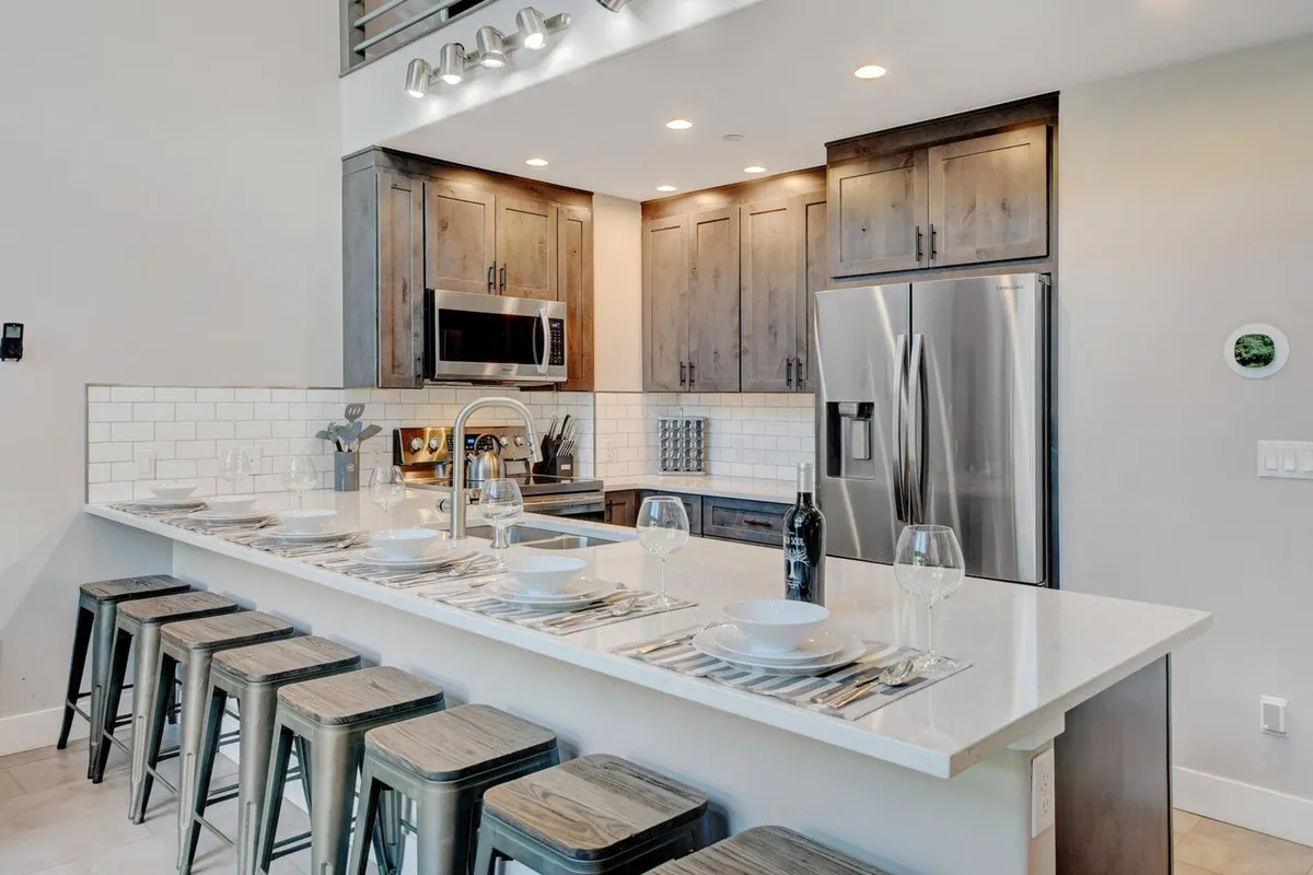 This image showcases a bright, contemporary kitchen with a central island, sleek stainless steel appliances, white cabinetry, and bar stools positioned at the breakfast bar. The open layout invites guests to gather, cook, and converse, with room for both culinary activity and casual dining.