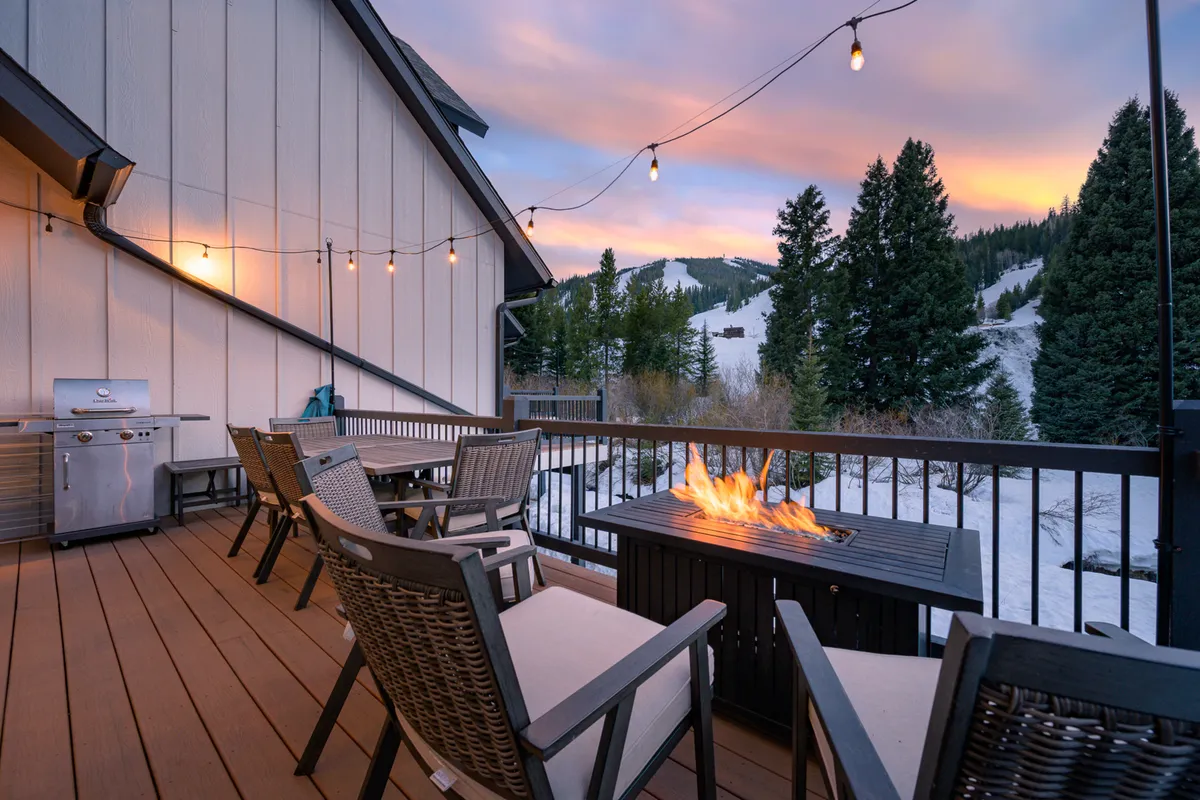 An outdoor dining set and fire pit sit beneath twinkling string lights on a spacious deck, with mountains providing a majestic backdrop. The scene is set for evening gatherings, al fresco dining, or simply enjoying the warmth of the fire with loved ones.