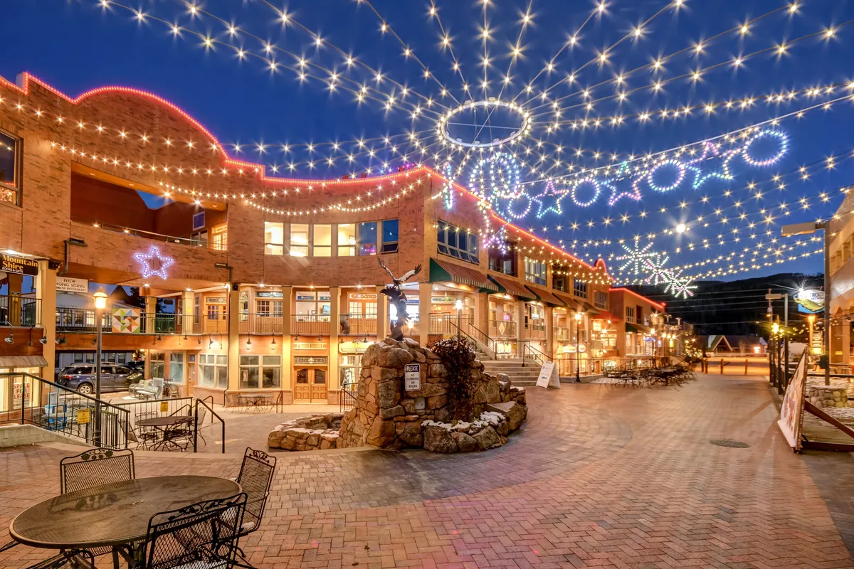 A charming town scene at dusk, illuminated by twinkling overhead lights and decorative stars, with a stone fountain and bustling storefronts. People stroll, dine outdoors, and enjoy the festive atmosphere that characterizes the area year-round.
