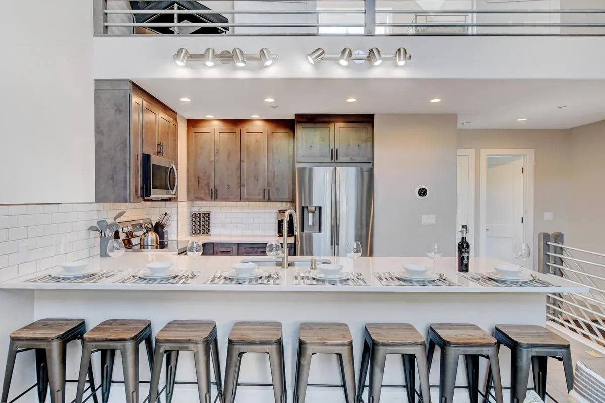 Here, the kitchen’s island, flanked by bar stools and surrounded by stainless steel appliances and warm wooden cabinetry, creates a welcoming space for cooking and socializing. The room’s design encourages guests to gather for shared meals or cooking experiences, with a clear view of the communal atmosphere.